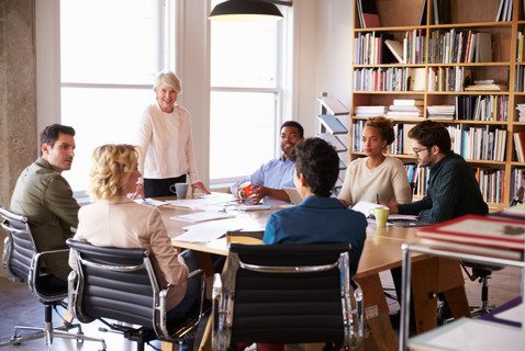 Eine mittelalte Frau steht an einem Tisch vor einer Gruppe sitzender, jüngerer Personen