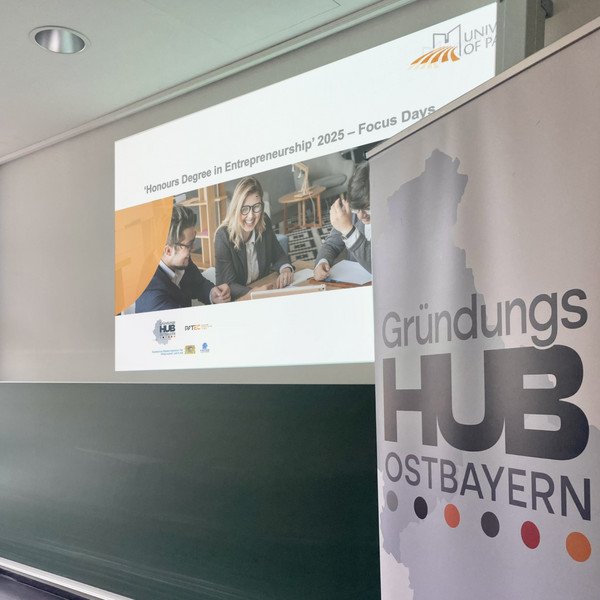 Im Vordergrund ist ein Roll-up zu sehen, auf dem das Logo des Gründungshubs Ostbayern gedruckt ist. Im Hintergrund ist eine Powerpoint-Folie mit einem Bild und der Aufschrift "Honours Degree in Entrepreneurship" und "Focus Days" abgebildet.